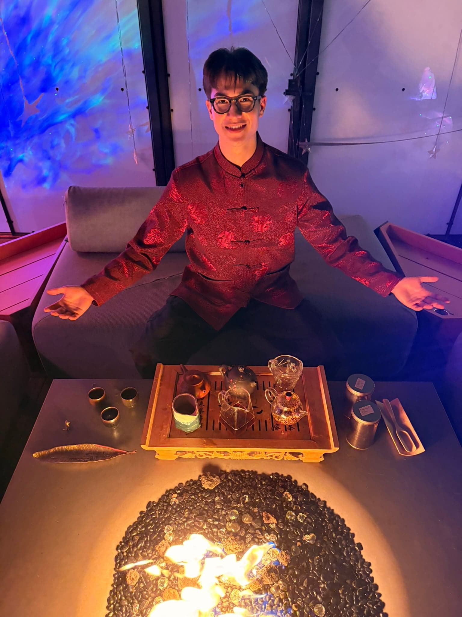 Ethan Holtzman seated at a tea ceremony in a red silk jacket, warm light around him